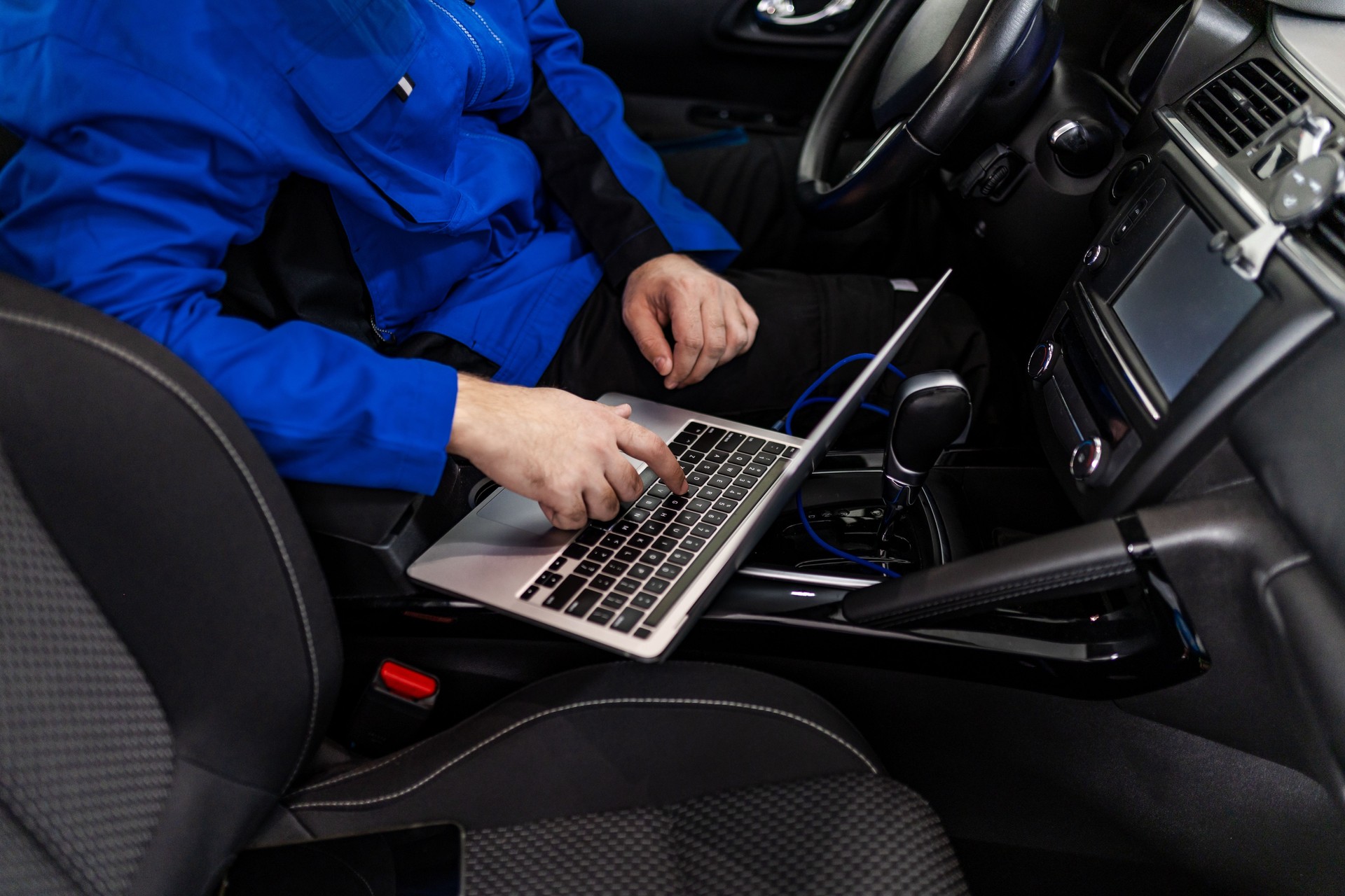 A technician works on a laptop inside a modern vehicle in an automotive workshop, focusing on diagnostics and repairs during daylight hours